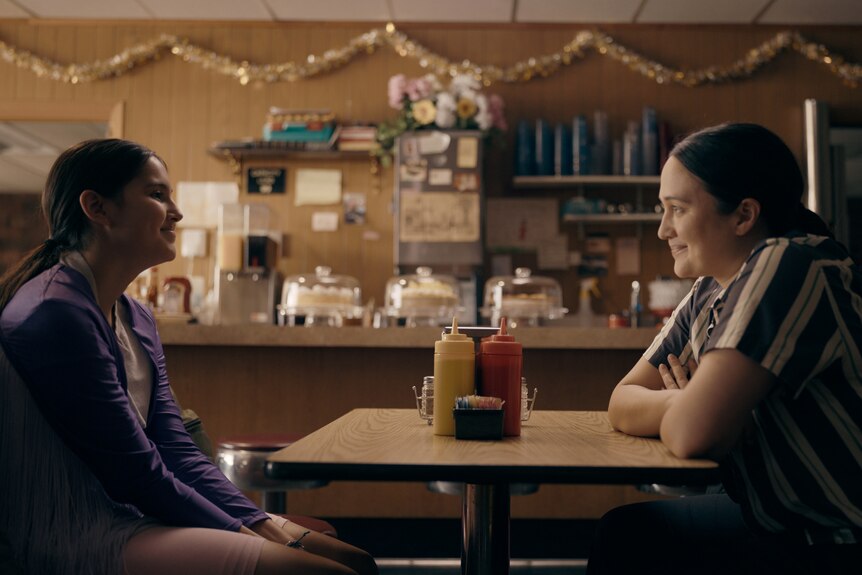 A woman and a teenage girl sit across from each other at a diner table, smiling.
