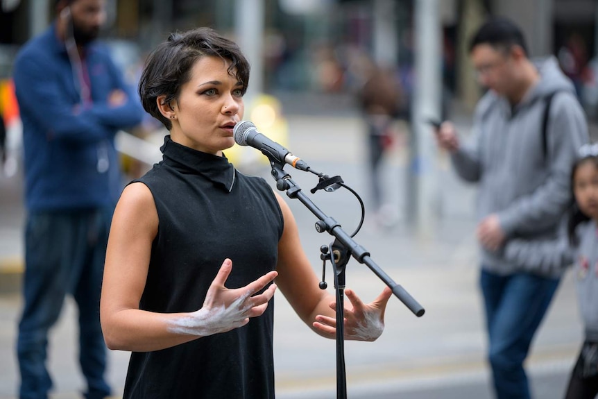 Young woman with short dark hair singing into microphone outdoors, passersby behind her.