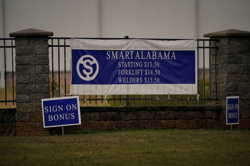 a blue and white banner tied to a fence advertises wages for workers at Smart Alabama