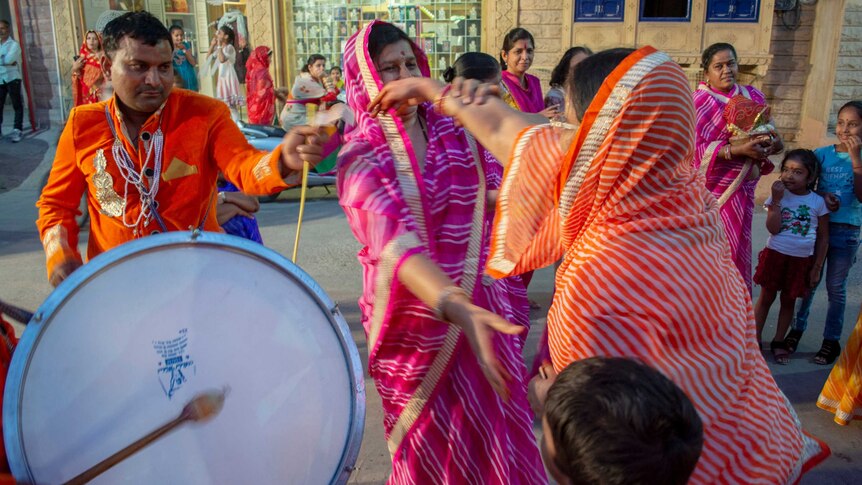 On the streets of India a group of women and men in brightly coloured clothes, of pink and orange dance to the beat of a drum.