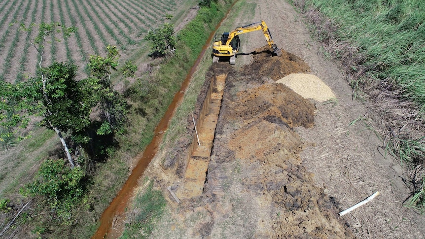 A mechanical digger digs out a long trench in a paddock.