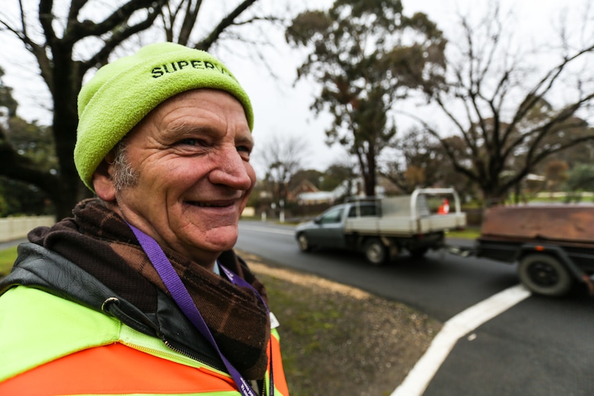 Close up of Ray Stevenson's face. He wears a fluorescent beanie with word 'supervisor' embroidered. A car and trailer travel by.