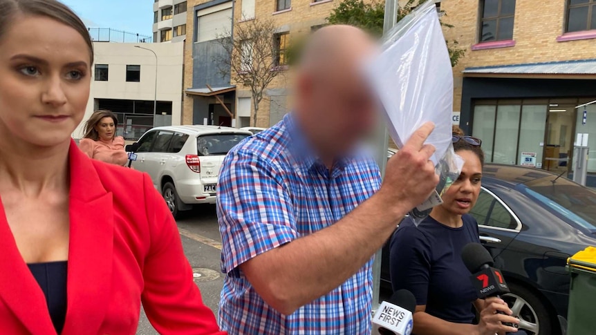 A middle-aged man puts a plastic folder of papers in front of his face as he walks past reporters.