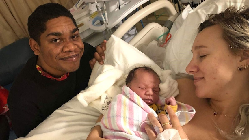 Man kneels next to a woman in a hospital bed, both holding their new born son.