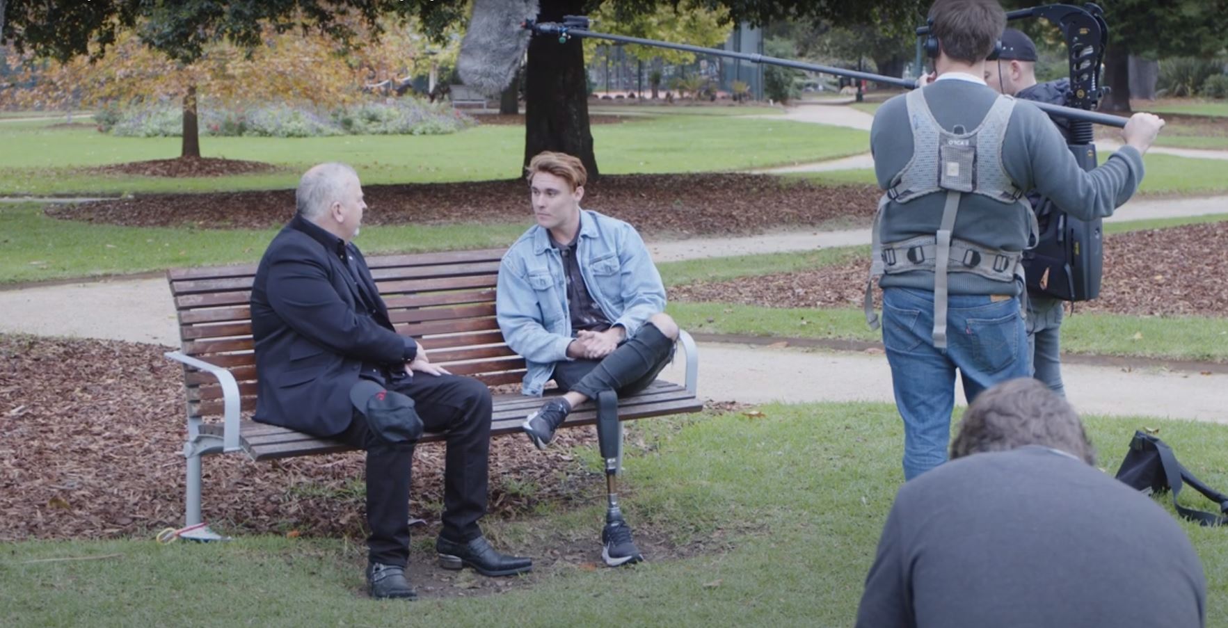 Joey Fry sitting on a park bench talking to a man, while a camera and sound team captured the interaction, date unknown.