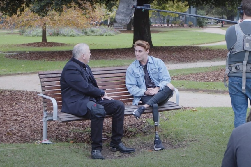 Joey Fry sitting on a park bench talking to a man, while a camera and sound team captured the interaction, date unknown.