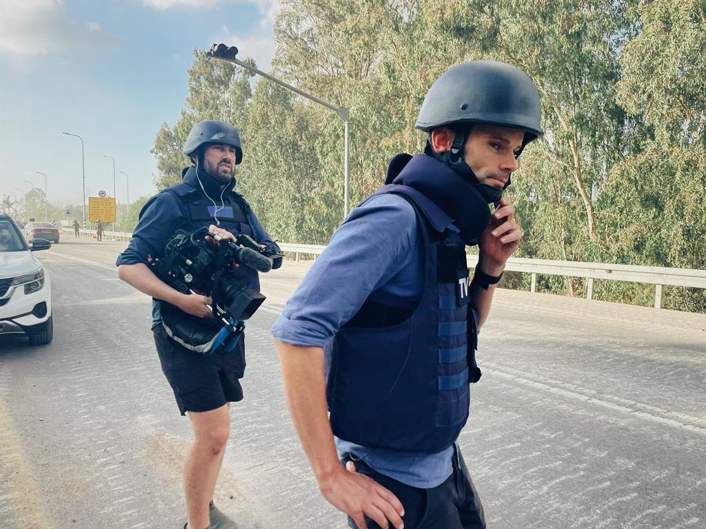 Two men in flak jackets and helmets in a street, one holding a TV camera, both looking down street assessing the situation.