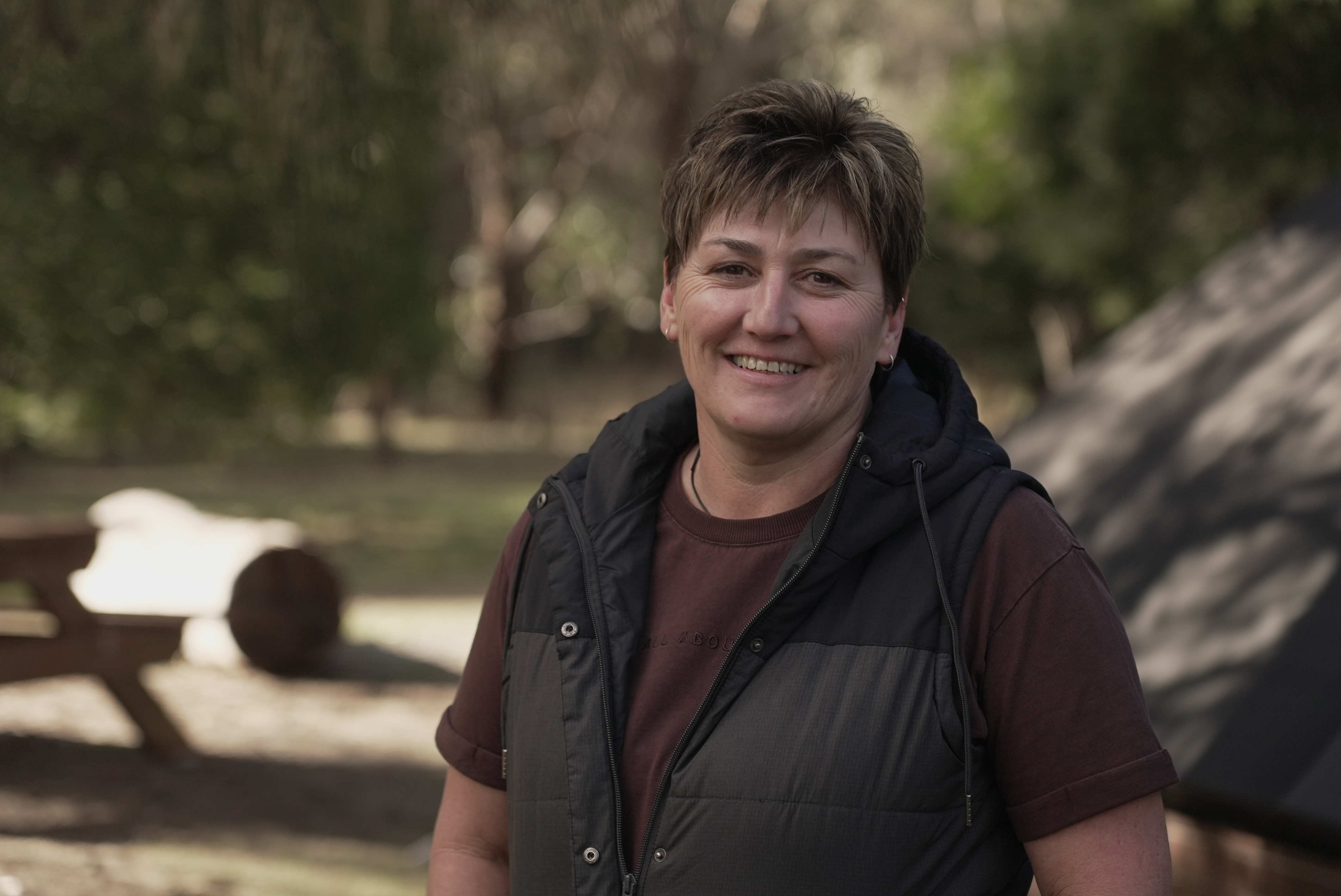 A woman with a warm vest smiles at the camera while standing in a campground.
