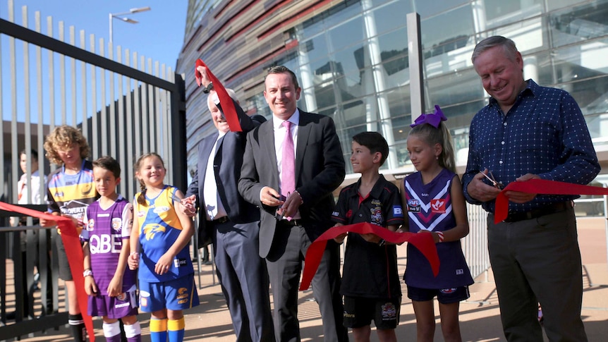 Three men and five kids, dressed in different team colours, cuts a large ribbon.