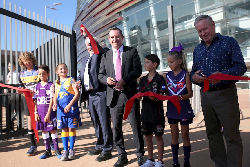 Three men and five kids, dressed in different team colours, cuts a large ribbon.
