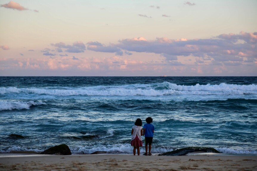 Two little kids stand on the beach looking out at the sunset 