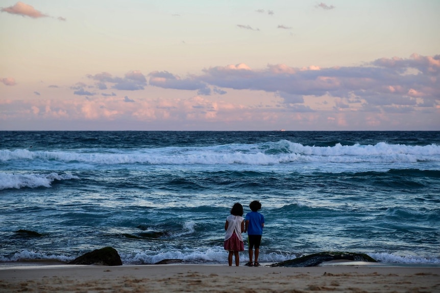 Two little kids stand on the beach looking out at the sunset 