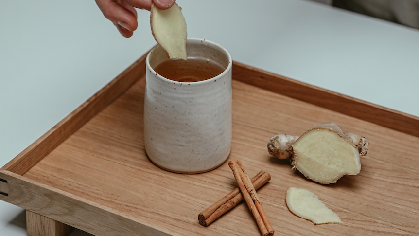 A person places a slice of ginger into a cup of tea, which sits on a tray next to cinnamon and more ginger. 