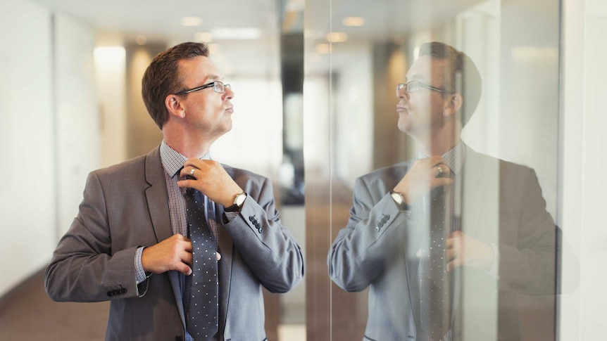A man in a grey suit fixes his tie in the reflection on a glass wall with a haughty look on his face.