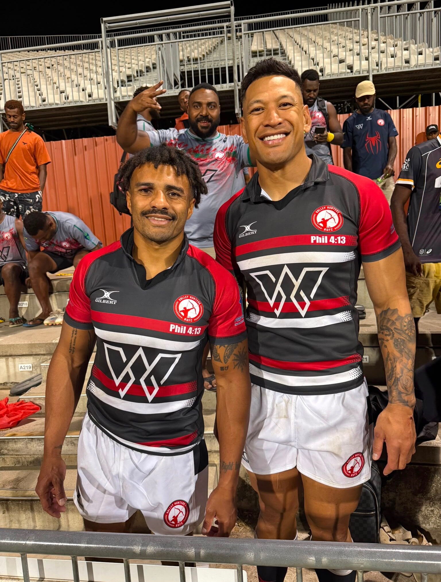 Papua New Guinean man and a taller Tongan man stand together wearing the same uniforms on stadium seats with people behind them