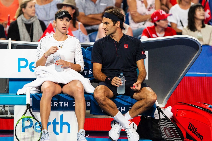 Roger Federer sits next to Belinda Bencic and they drink water during a drinks break on the tennis court.