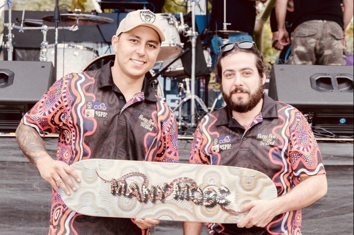 Two men smile as they both hold a skateboard that says "Many mobs". They are wearing shirts emblazoned with Indigenous art.