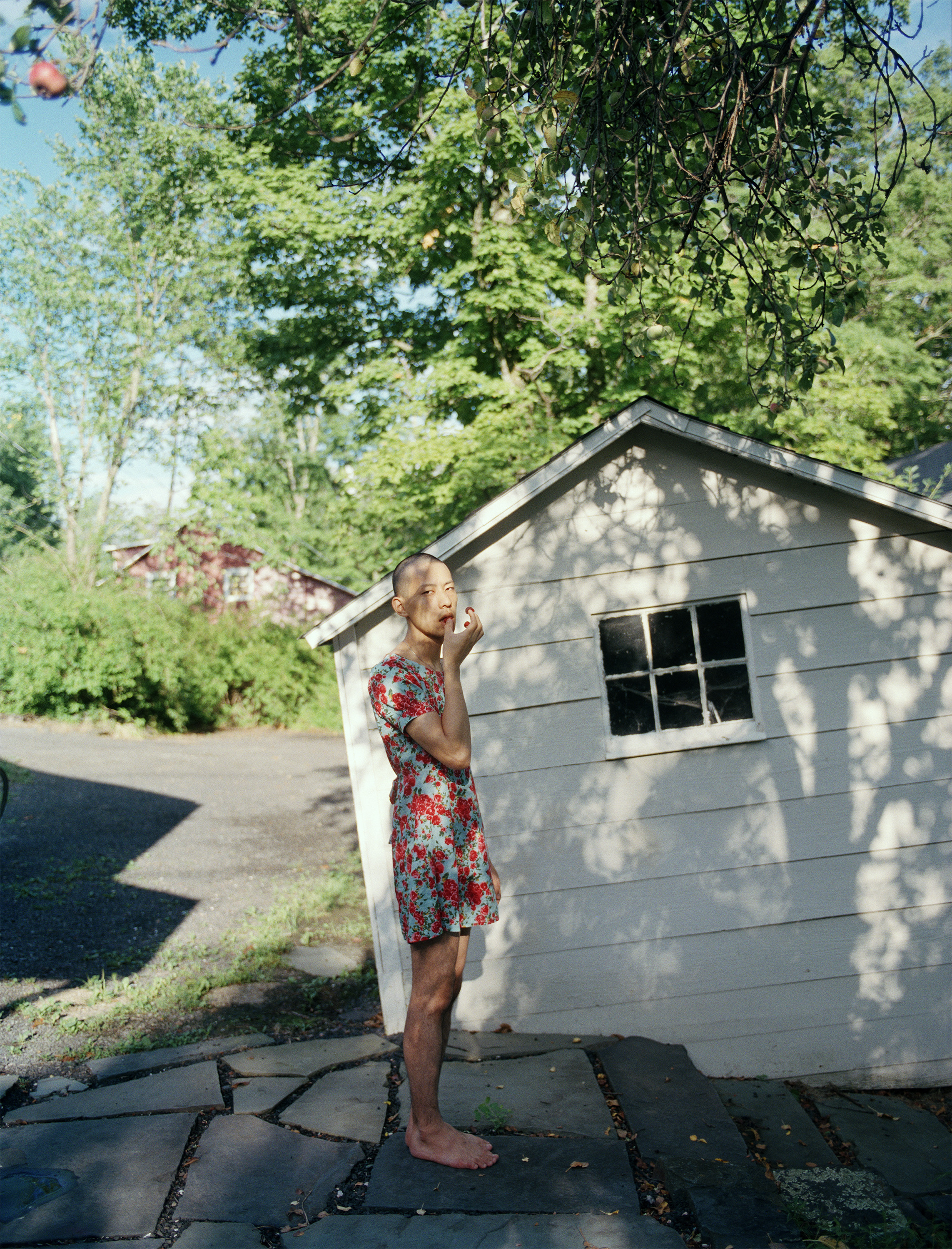 Moro, a Japanese man with a close-shaved head wears a blue and red floral dress, licking his fingers beside a white shed