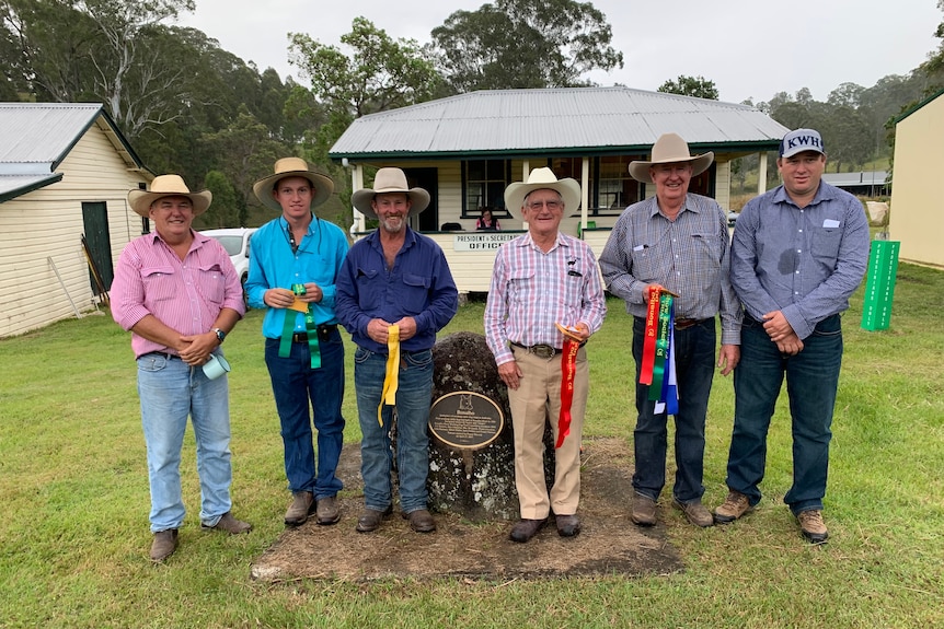 Six men wearing hats and holding ribbons stand three each side of a stone featuring a plaque with a working dog.