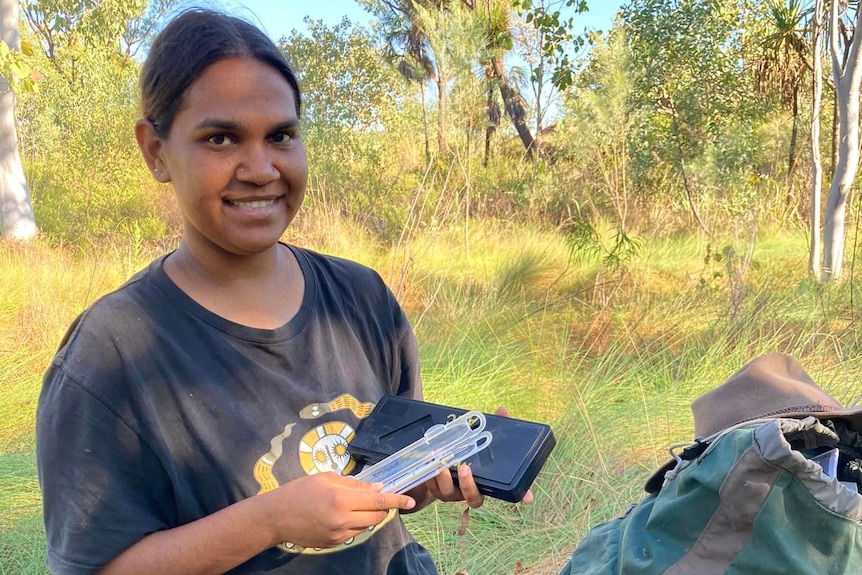 A young Aboriginal woman in a black t-shirt holds up measuring devices in front of a bush setting.