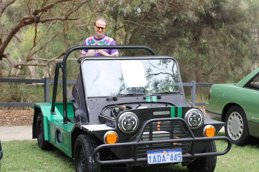 A man wearing a t-shirt and sunglasses stands in the back of a 70s Moke.