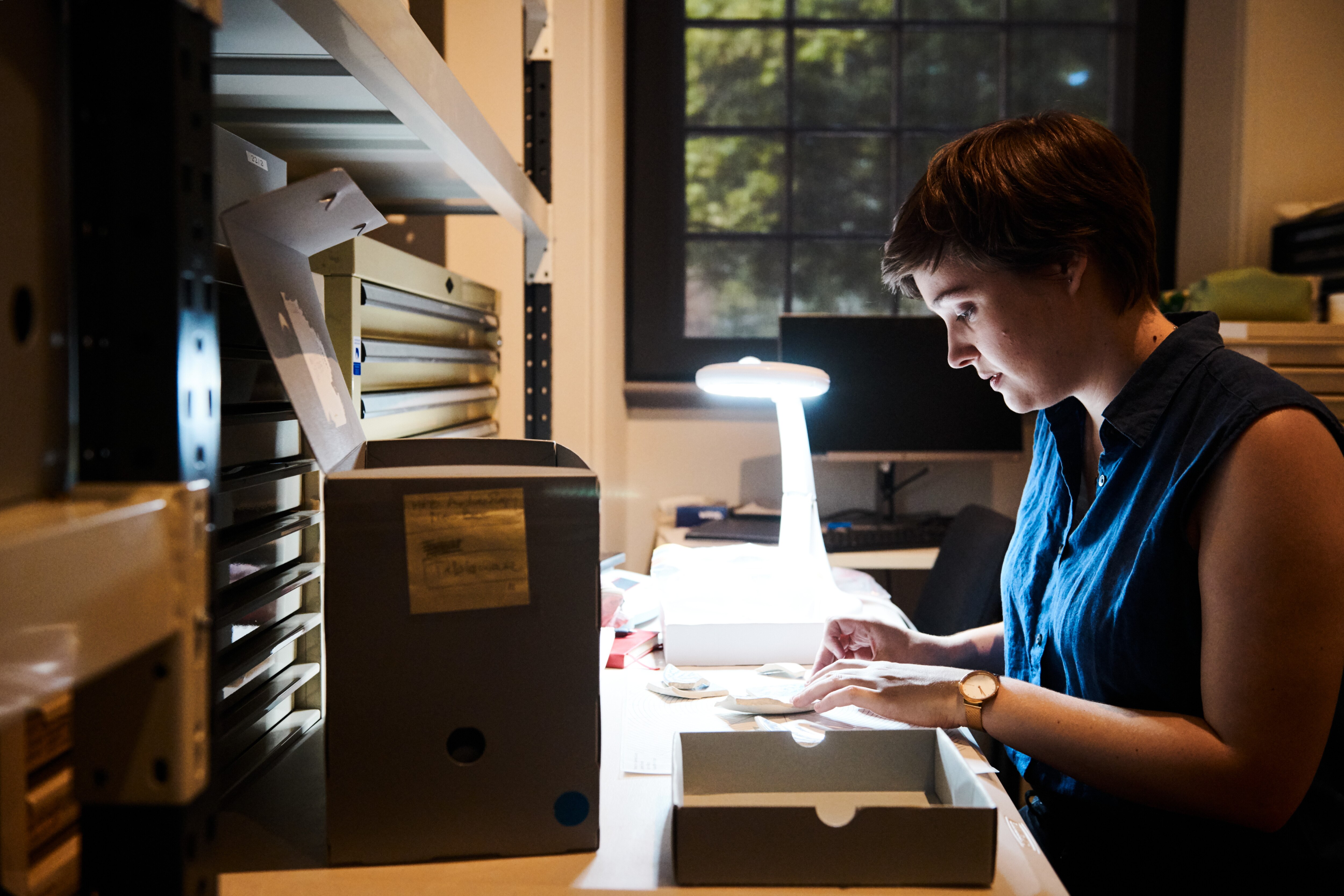 A woman with short brown hair and a blue shirt sits in a office studying a sample under a light.