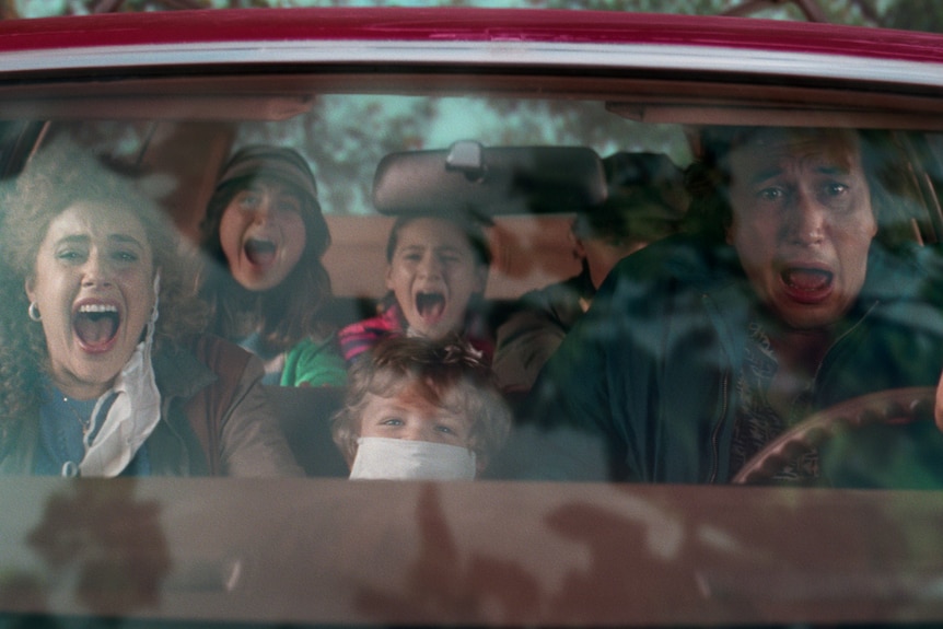 A family in 80s dress - including a teenage son, a teenage daughter, a young boy, and a pre-teen girl - scream in a car.