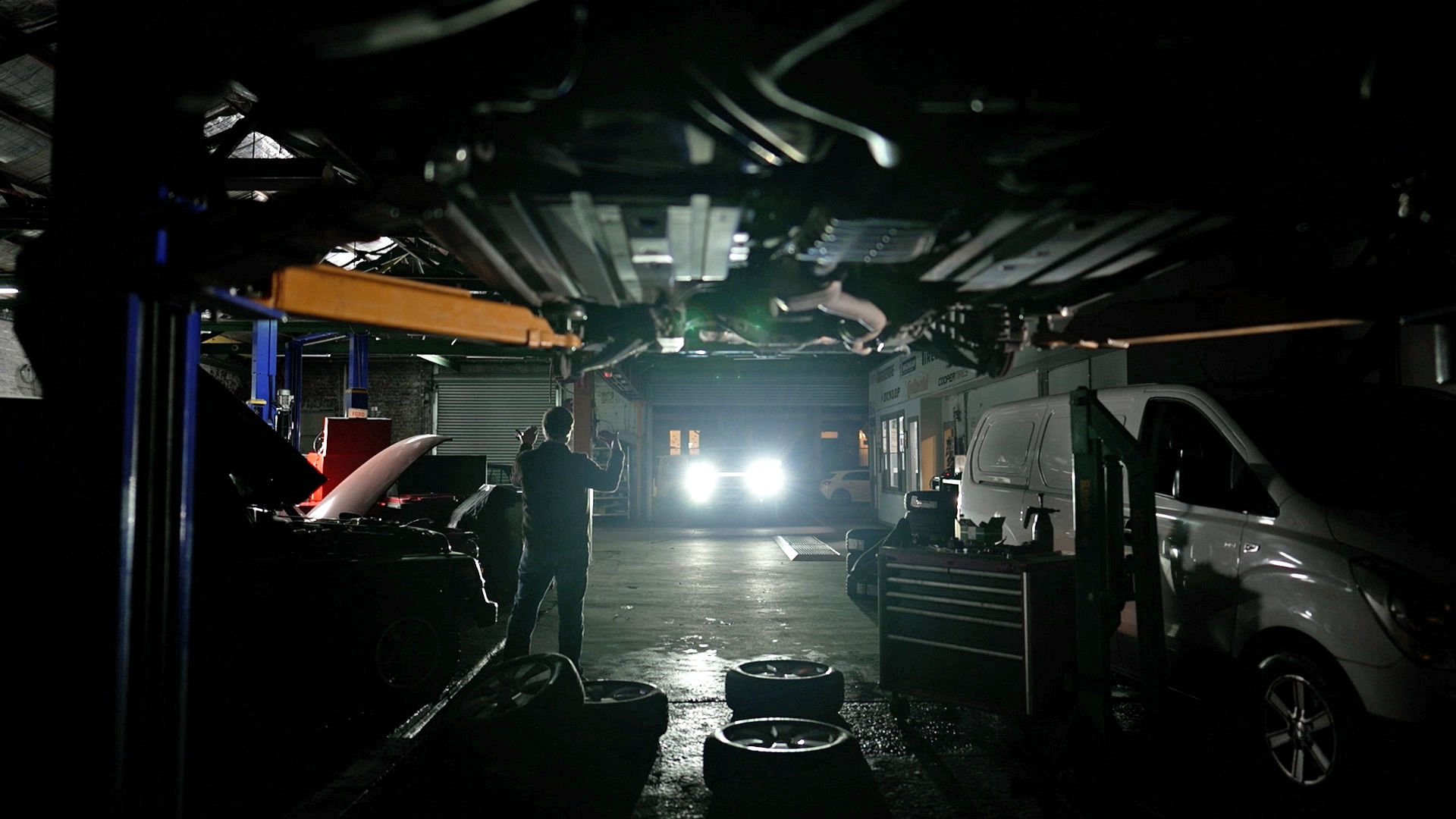 暗い整備士のガレージ。部屋に入ってくる車のヘッドライトに向かって身振りで示す男性。クレーンの上に車が乗っています。