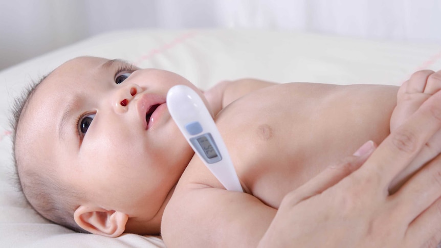 a very young baby lies on a bed and is having his temperature taken using a simple armpit digital thermometer