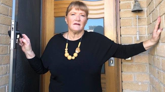 A blonde woman wearing a black dress and a gold chain stands in a doorway looking angry.