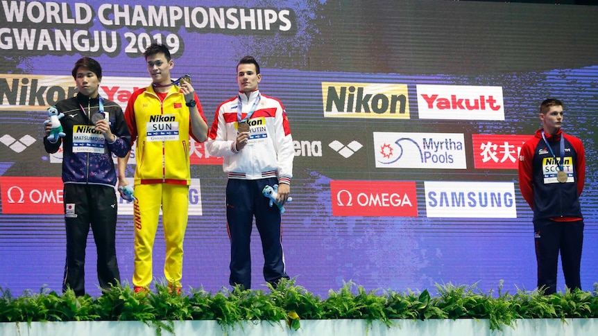 Three swimmers stand with their medals on a podium while a fourth is off to the side looking away