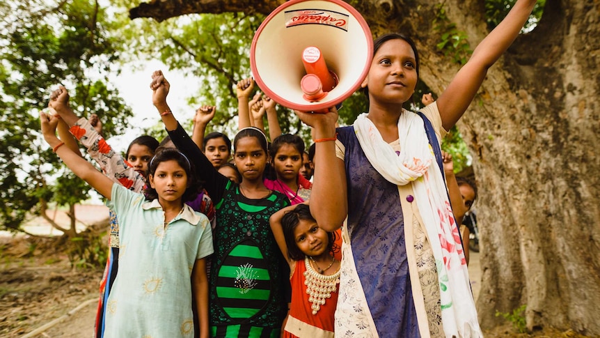 A group of girls in India standing together with their fists held in the air, and one holding a megaphone.