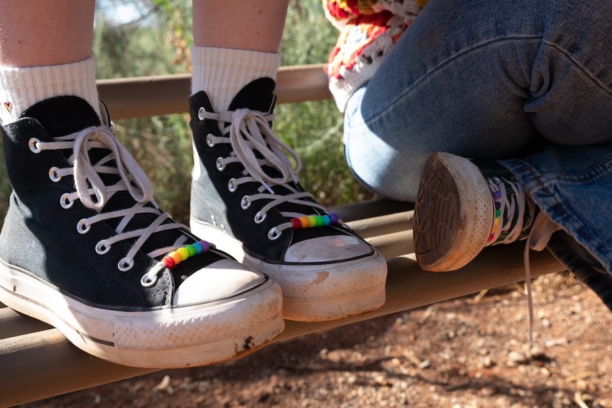 Close up of a pair of sneakers with rainbow beads threaded on their laces, standing on a bench.