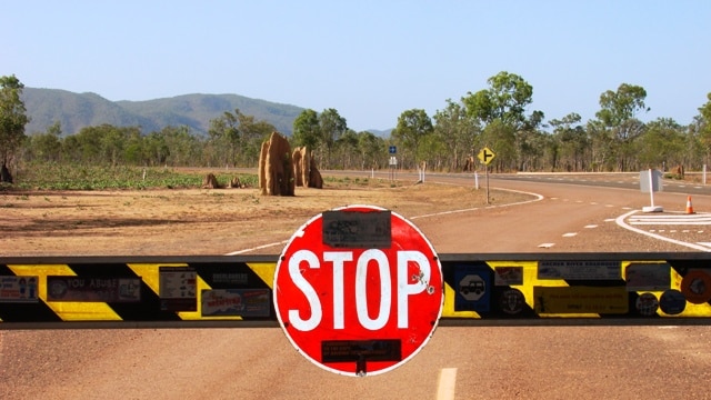 A stop sign is attached to a roadblock stopping vehicles, with anthills and bush landscape in the background