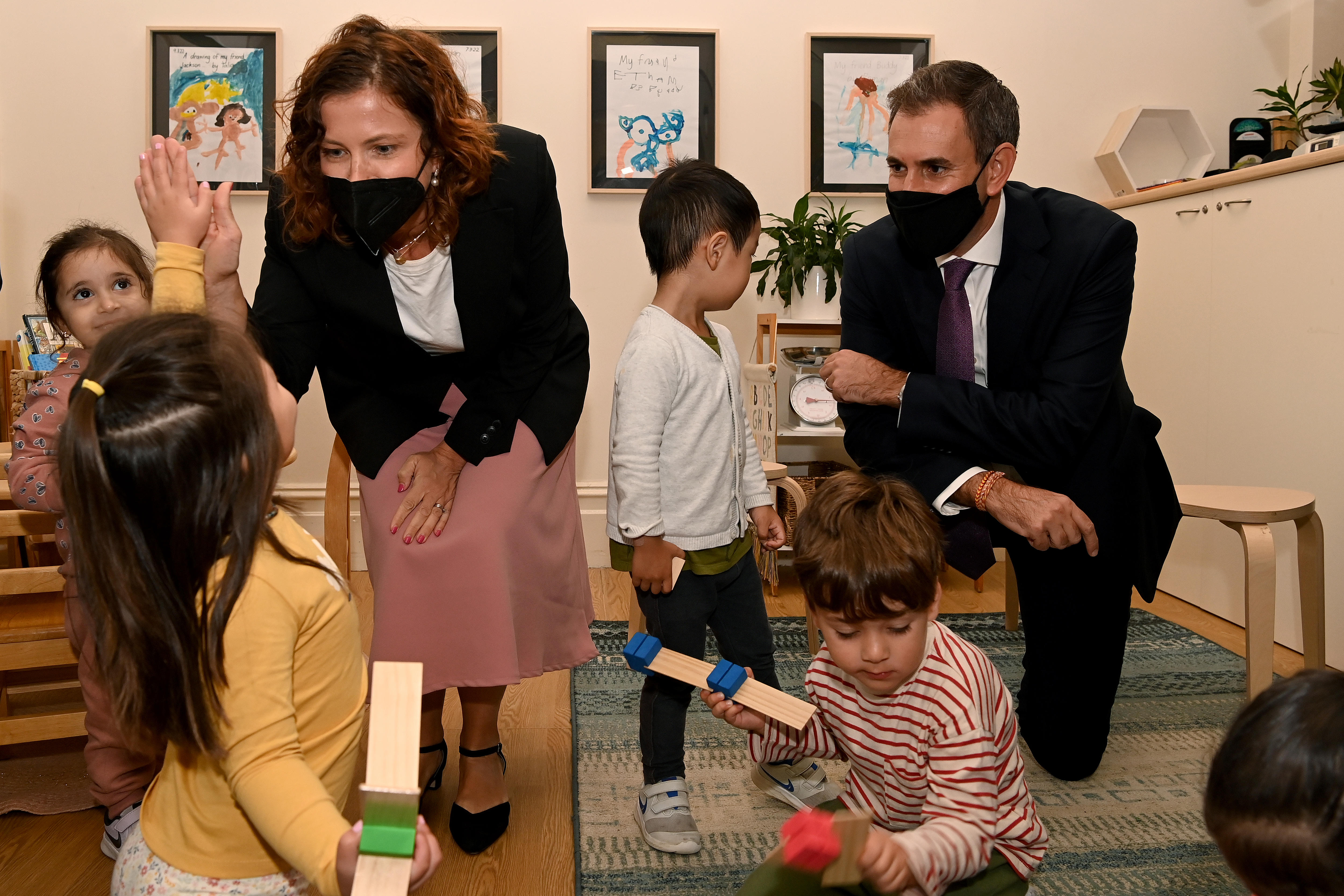 A man and a woman wearing face masks kneel down surrounded by young children and toys.