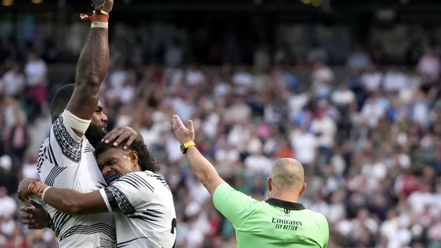 Two Fijian rugby players embrace happily in stadium full of spectators. 