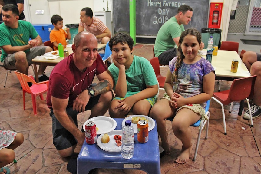 A man crouches down next to a kids size plastic table where his children sit, other men are in the background.