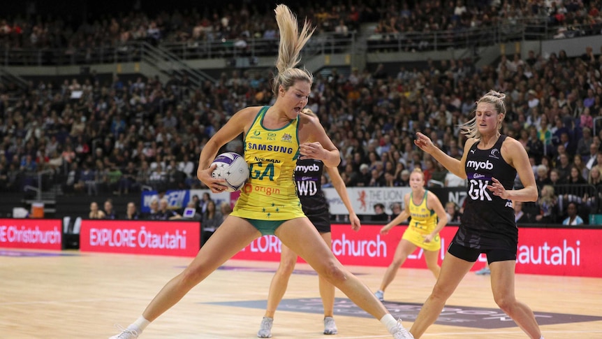 An Australian netball player holds the ball in her right had by her waist as she jumps in the air against New Zealand.