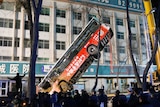 A bus is lifted up by a crane in front of a building with Chinese writing on it.