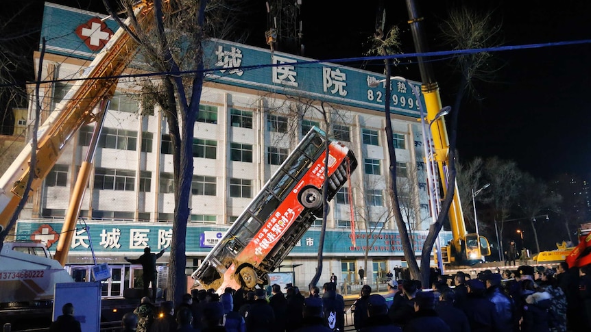 A bus is lifted up by a crane in front of a building with Chinese writing on it.