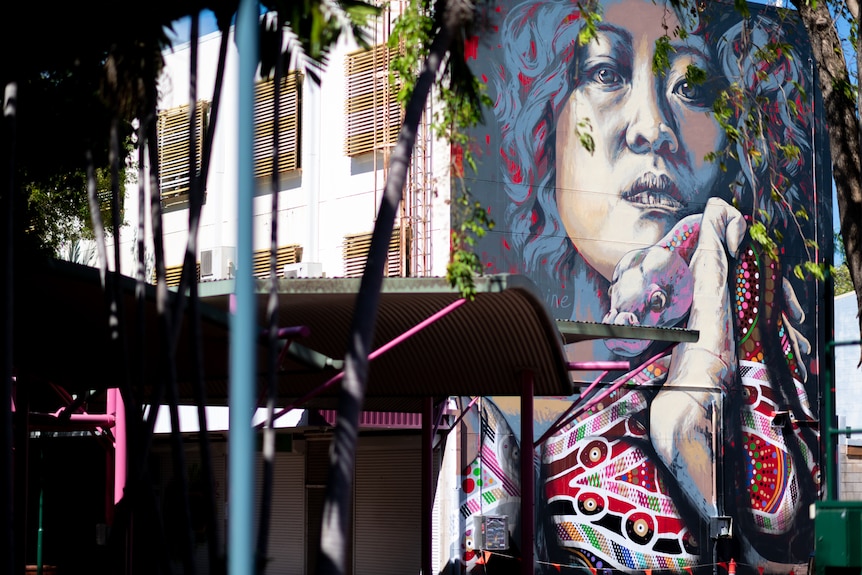 A mural of a woman's face painted on the side of a building in an outdoor mall, she is holding a snake near her face. 