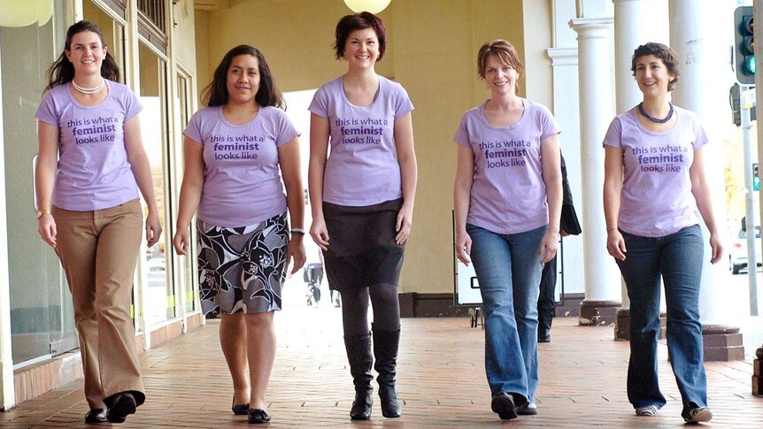 Five women walk along wearing shirts that say 'this is what a feminist looks like'.