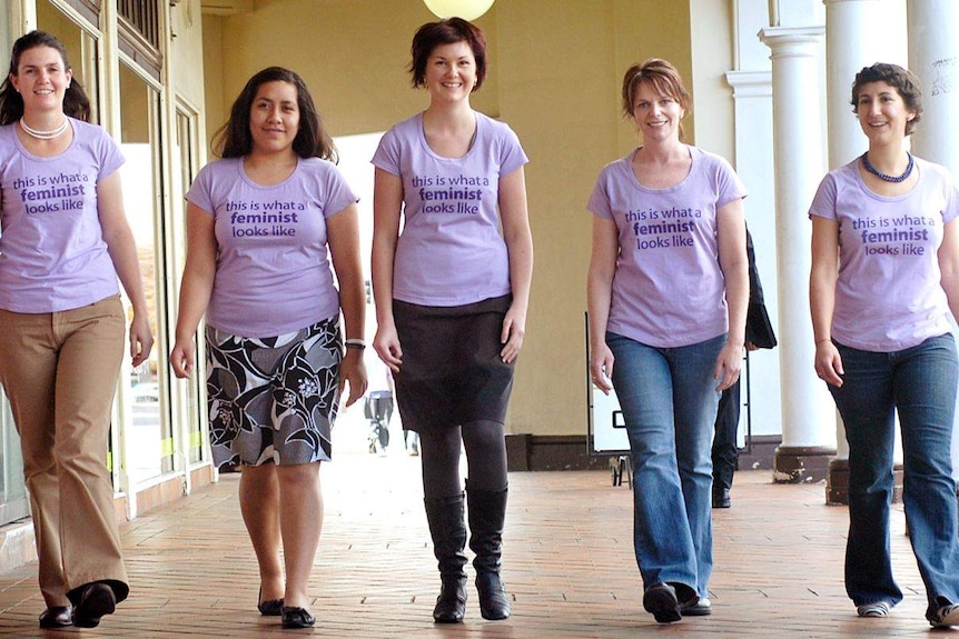 Five women walk along wearing shirts that say 'this is what a feminist looks like'.