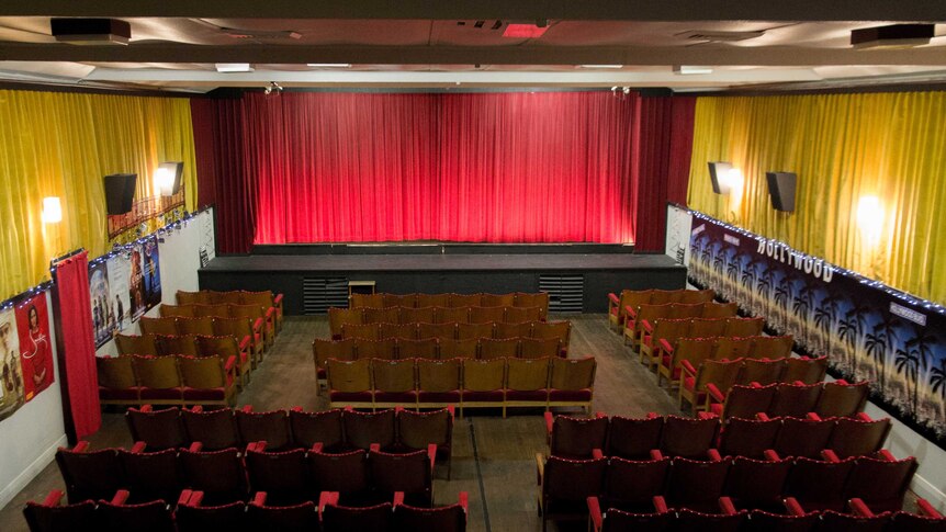 Rows of cinema seats face a screen covered by a red velvet curtain.