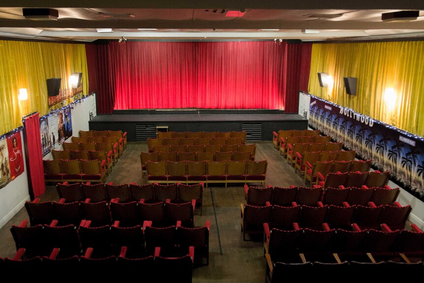 Rows of cinema seats face a screen covered by a red velvet curtain.