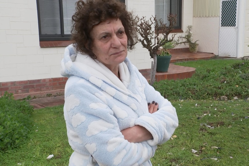 A woman in a dressing gown stands in front of her house with her arms folded