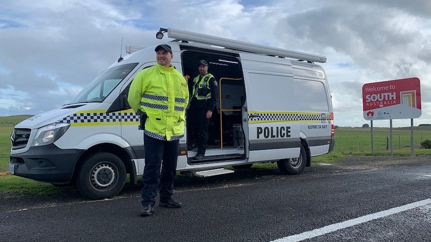 A policeman stands near a van and one inside a van next to a sign about entering South Australia.