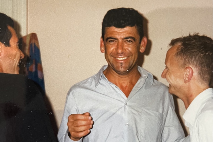 A man with dark hair stands smiling with friends at a social gathering. 