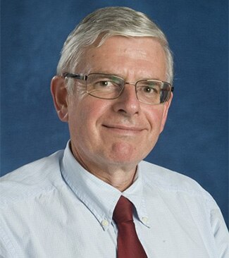 A man with grey hair and glasses in front of a blue backdrop. 
