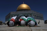 Muslim worshippers wrapped in Palestinian flags pray with heads down with gold dome of mosque in background.